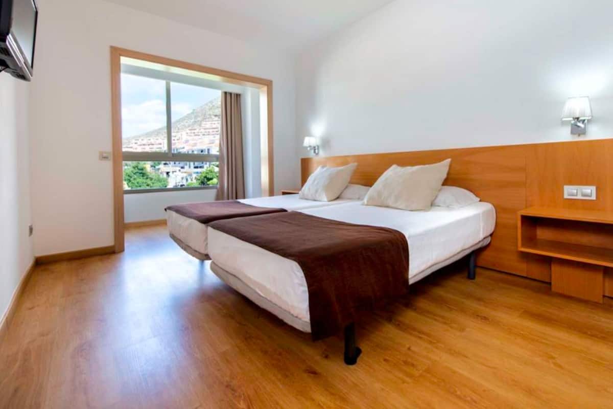 Habitación de hotel con cama, mesita de noche, lámpara y vista de la ciudad a través de un ventanal grande.
