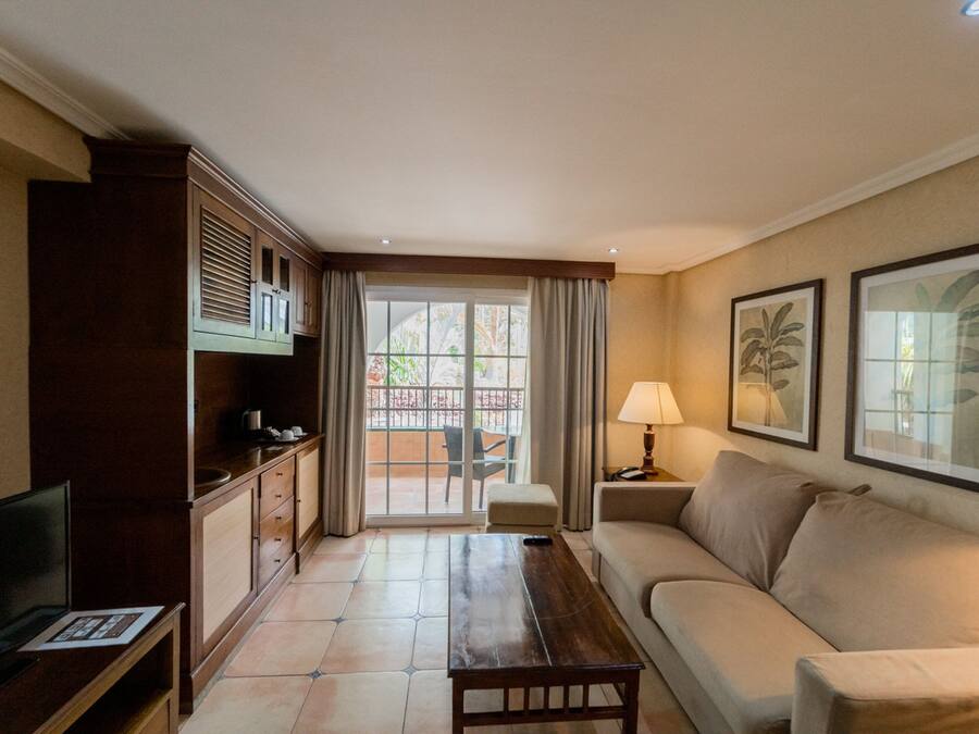 A hotel room with a sofa, a wooden coffee table, a TV, a desk with a lamp, and a sliding glass door leading to an outdoor area.