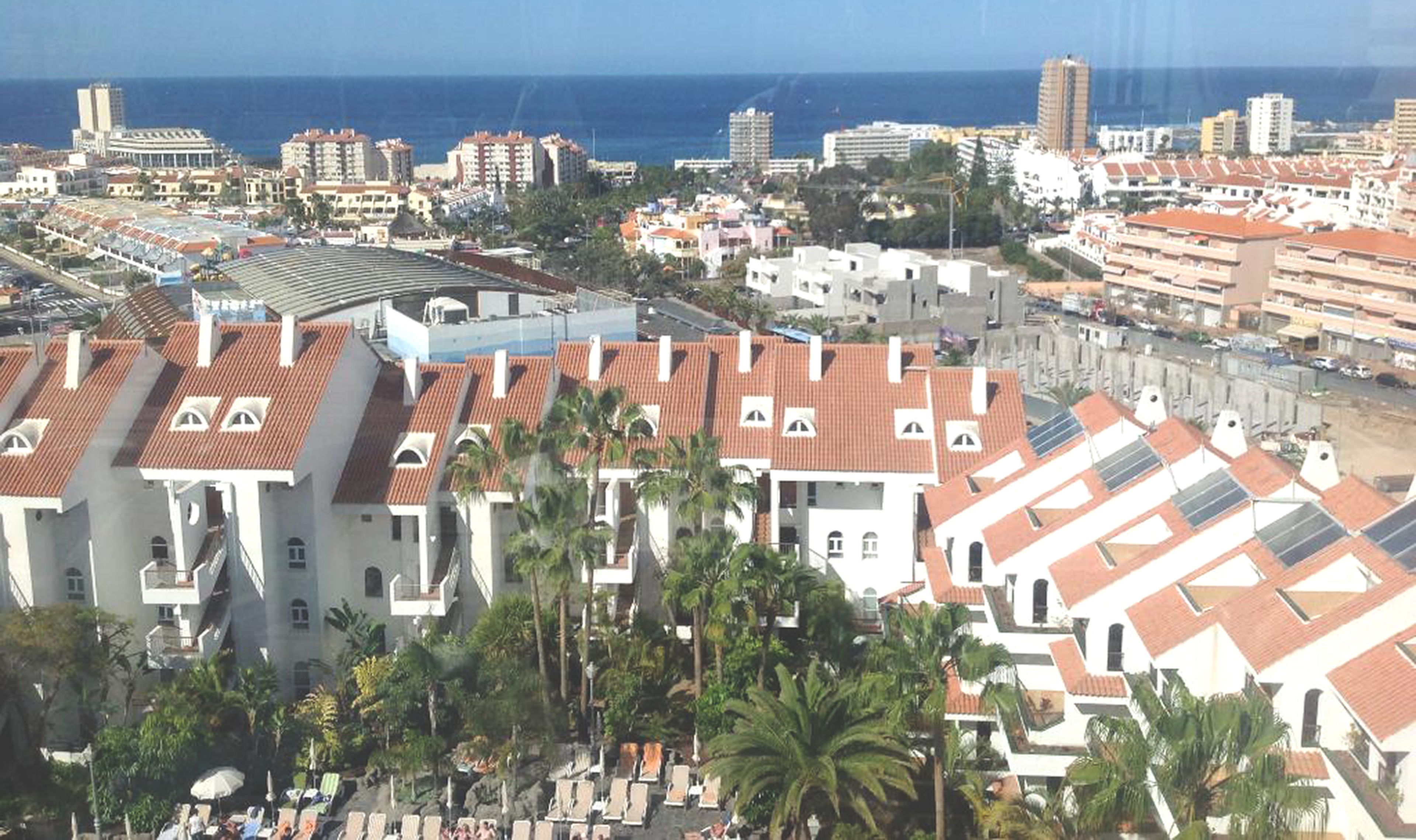 A coastal cityscape with a prominent hotel featuring terracotta roofs, palm trees, and a view of the sea.