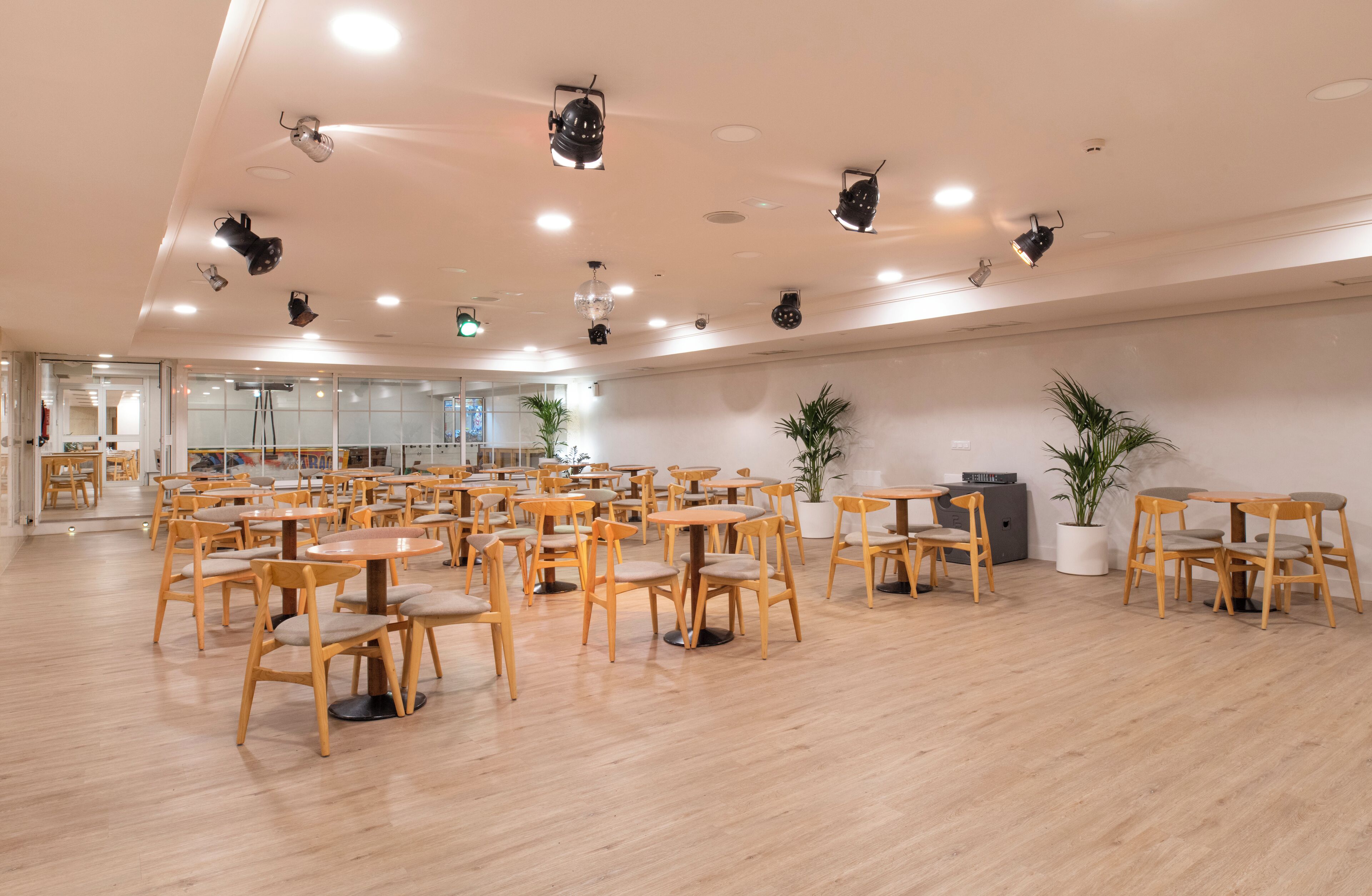 A spacious room with multiple tables and chairs, ceiling lights, and potted plants.