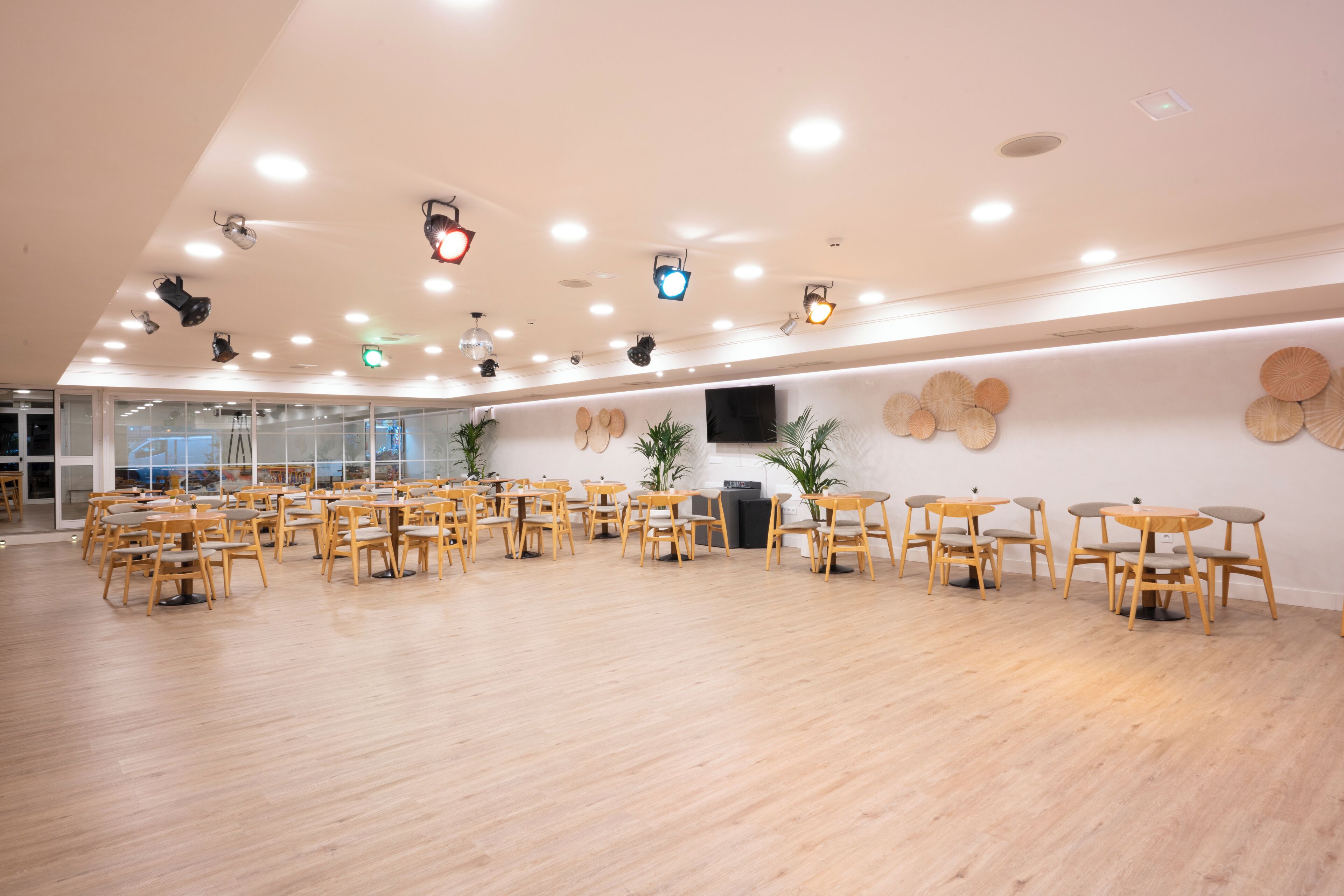 Une pièce spacieuse avec plusieurs tables et chaises en bois, un téléviseur à écran plat fixé au mur et plusieurs luminaires au plafond.
