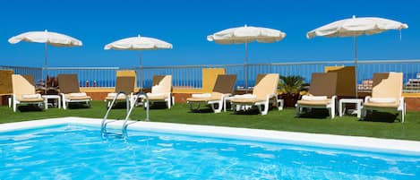 Zona de piscina con sillones de descanso y sombrillas, cielo azul despejado y vista al océano.