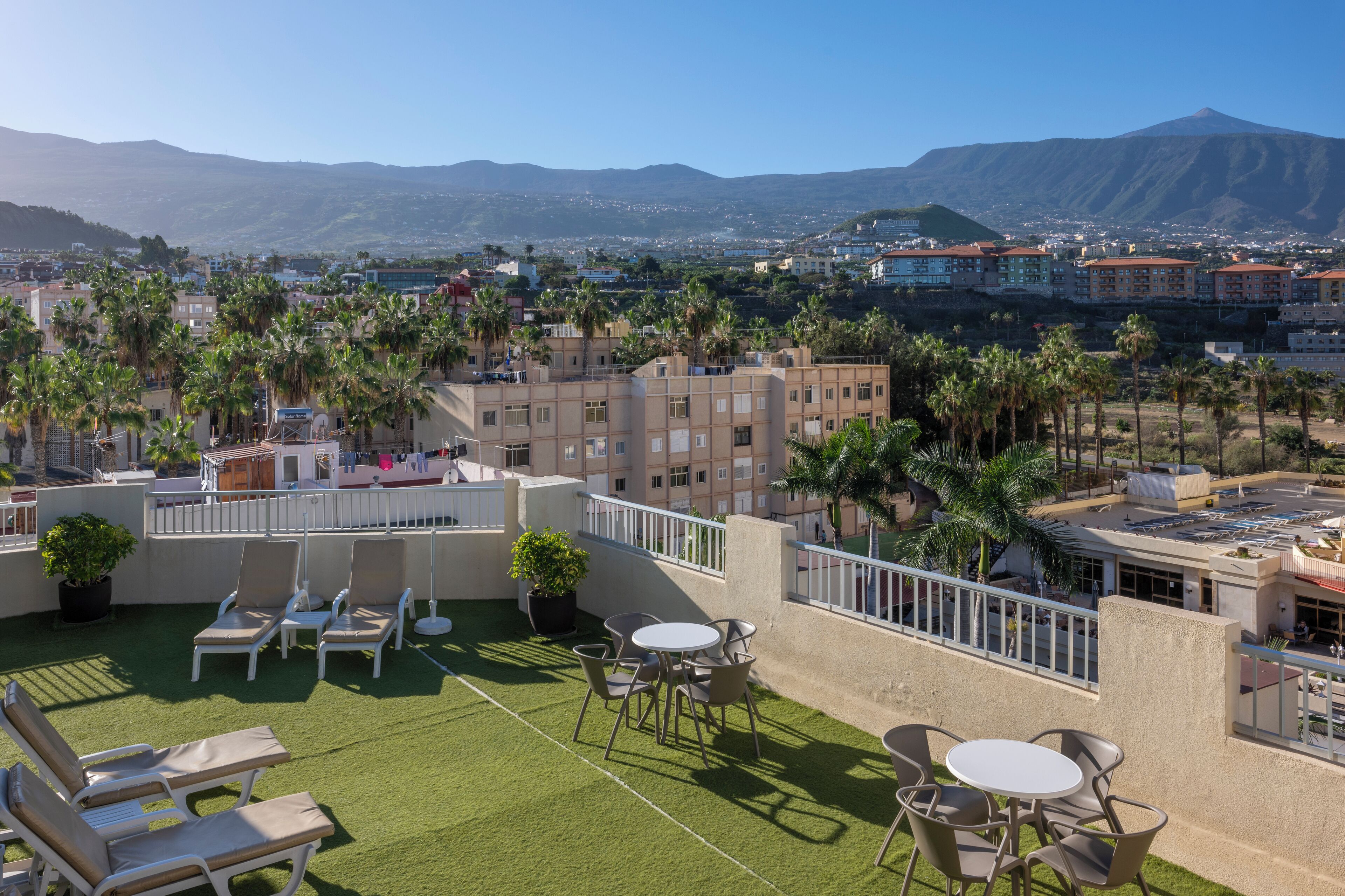 A rooftop terrace with artificial grass, lounge chairs, and tables, overlooking a cityscape with buildings and a mountain in the background.