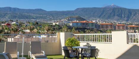 Eine Dachterrasse mit Liegestühlen und einem Tisch mit Blick auf eine Stadtlandschaft und ein Gebirge.