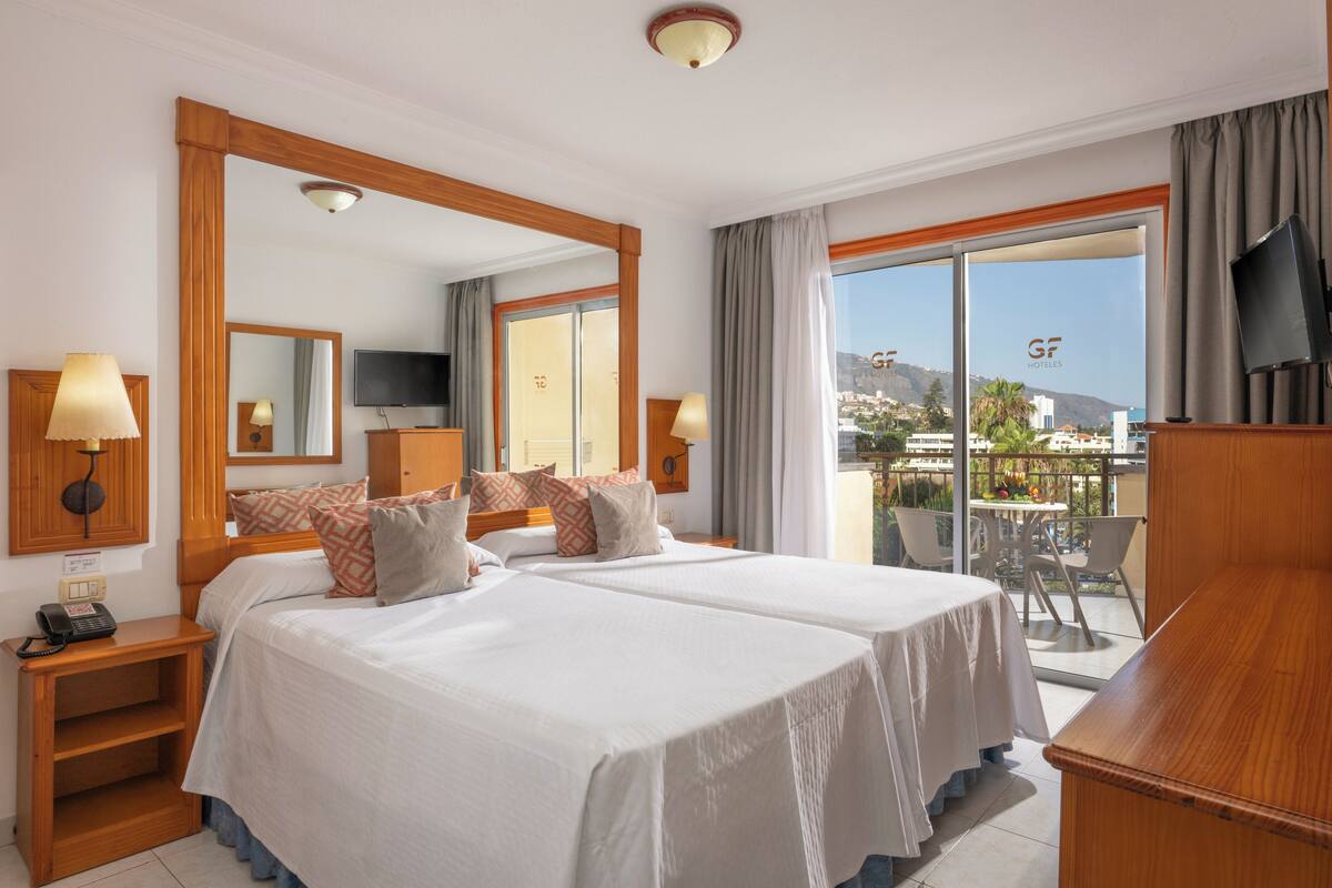 Habitación de hotel con dos camas, un balcón con mesa y sillas, televisión y un ventanal con vistas a la ciudad.