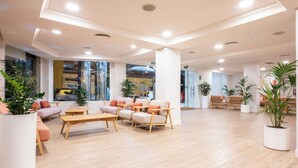 A spacious lobby with a modern design, featuring a seating area with sofas and chairs, a wooden coffee table, and potted plants.