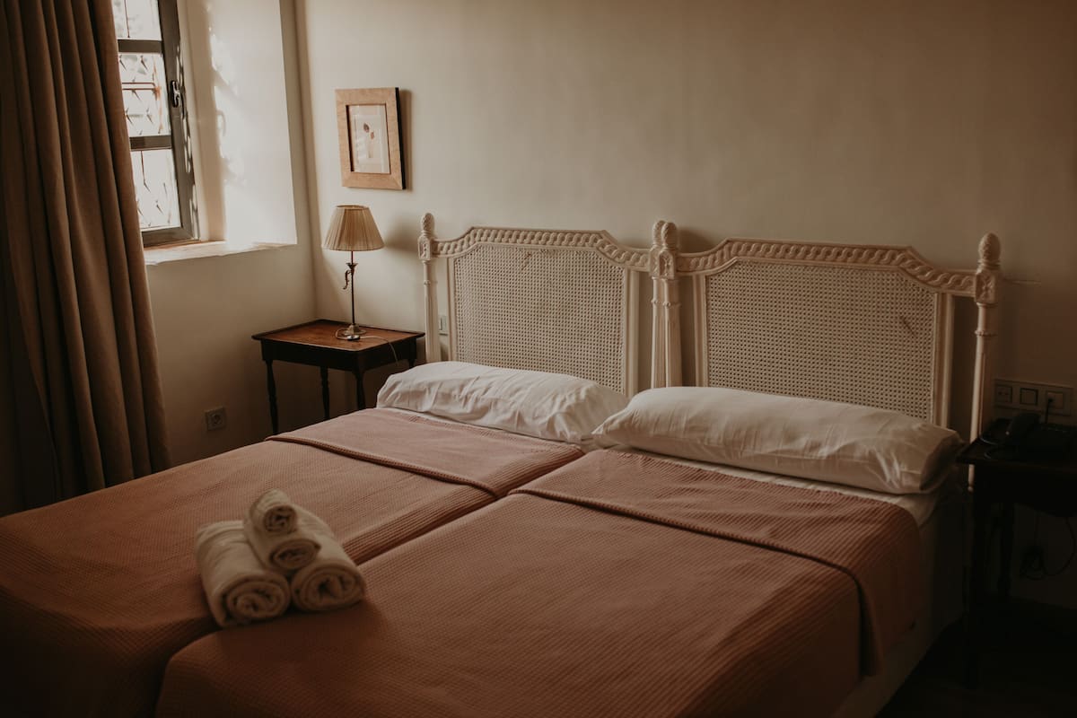 A double bed with a woven headboard, a bedside table with a lamp, and rolled towels on the bed.