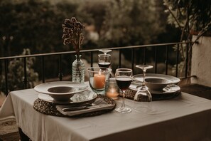 Una mesa preparada para una comida, con platos, vasos y una vela, al aire libre con barandilla y follaje de fondo.