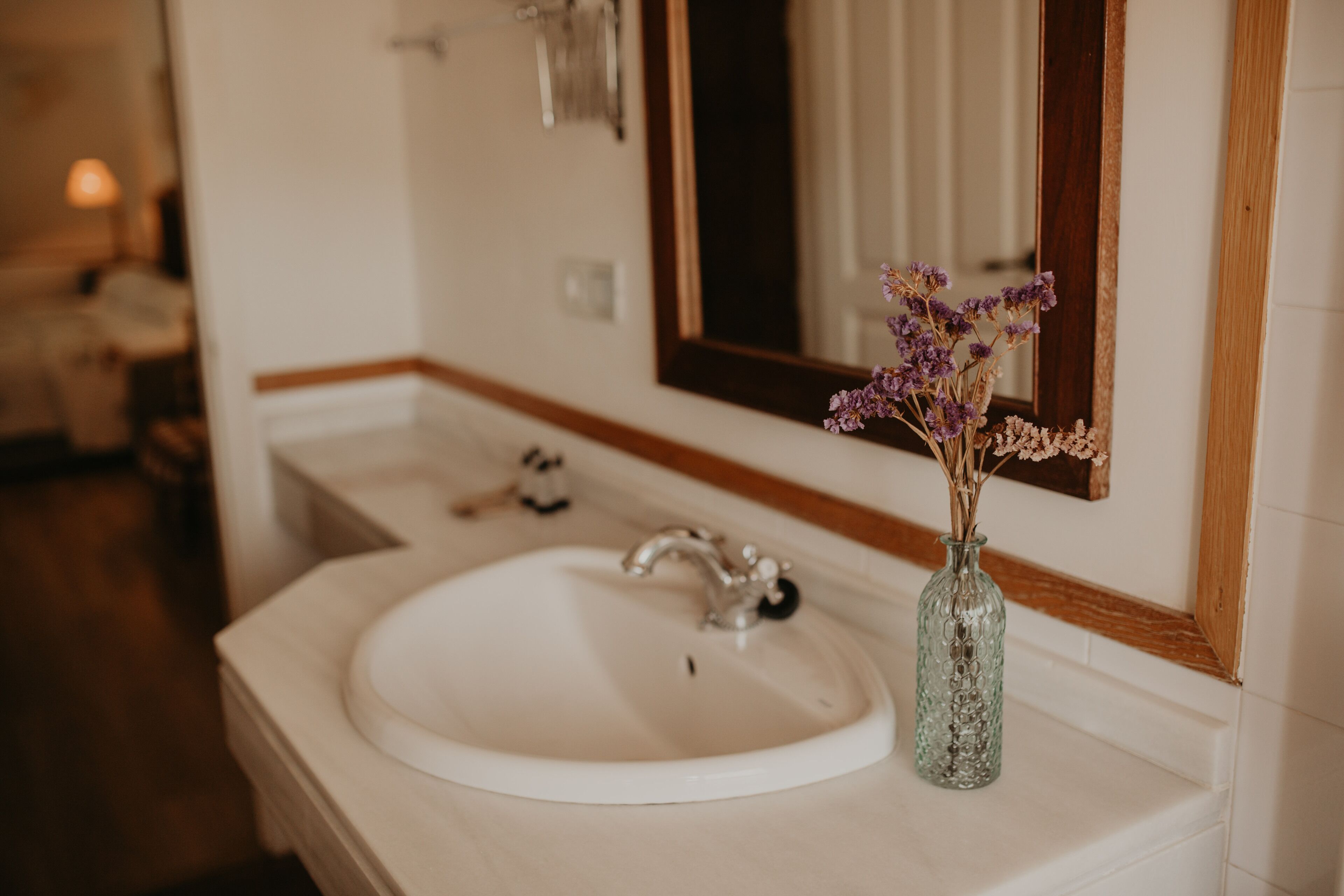 Un baño con un lavamanos blanco, un espejo y un florero con flores secas.