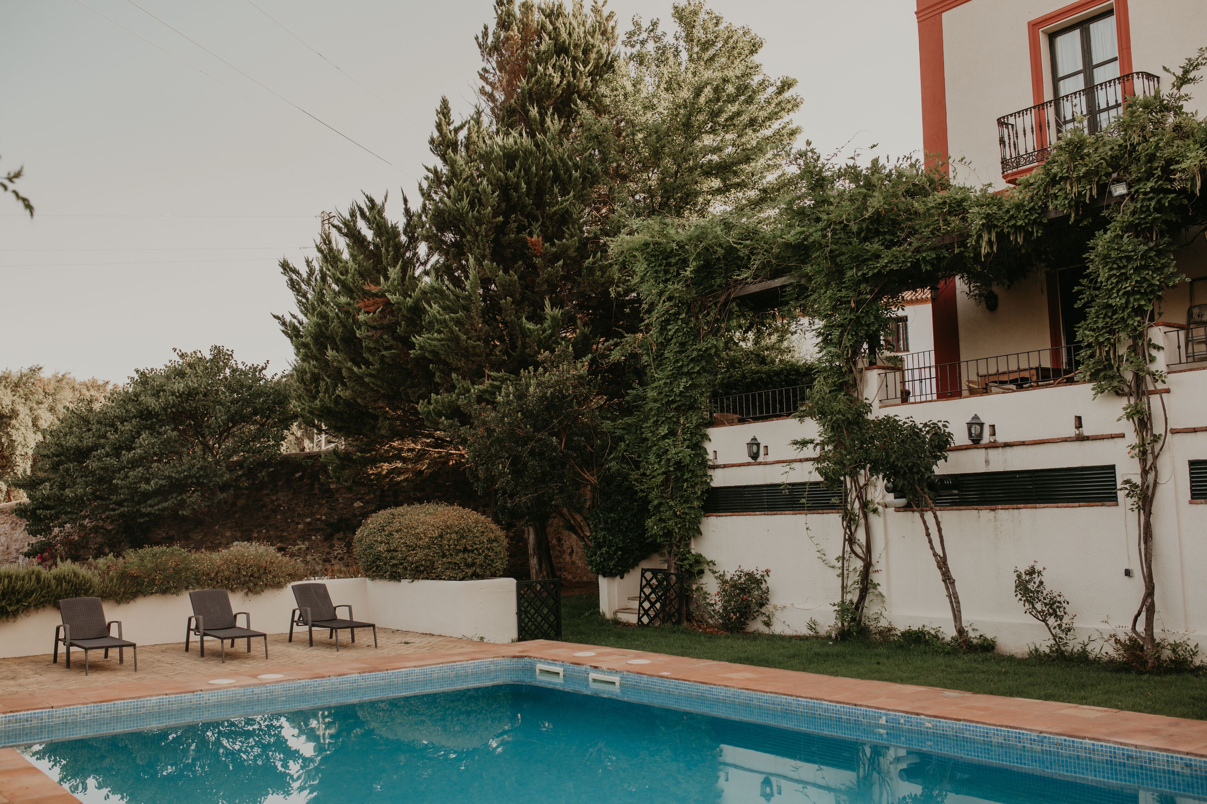 Una piscina con tres sillas de descanso, rodeada de vegetación y un edificio blanco con balcón.