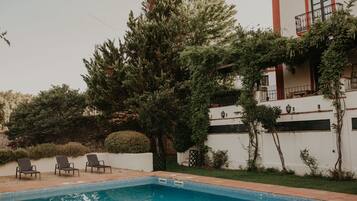 Una piscina con tres sillas de descanso, rodeada de vegetación y un edificio blanco con balcón.