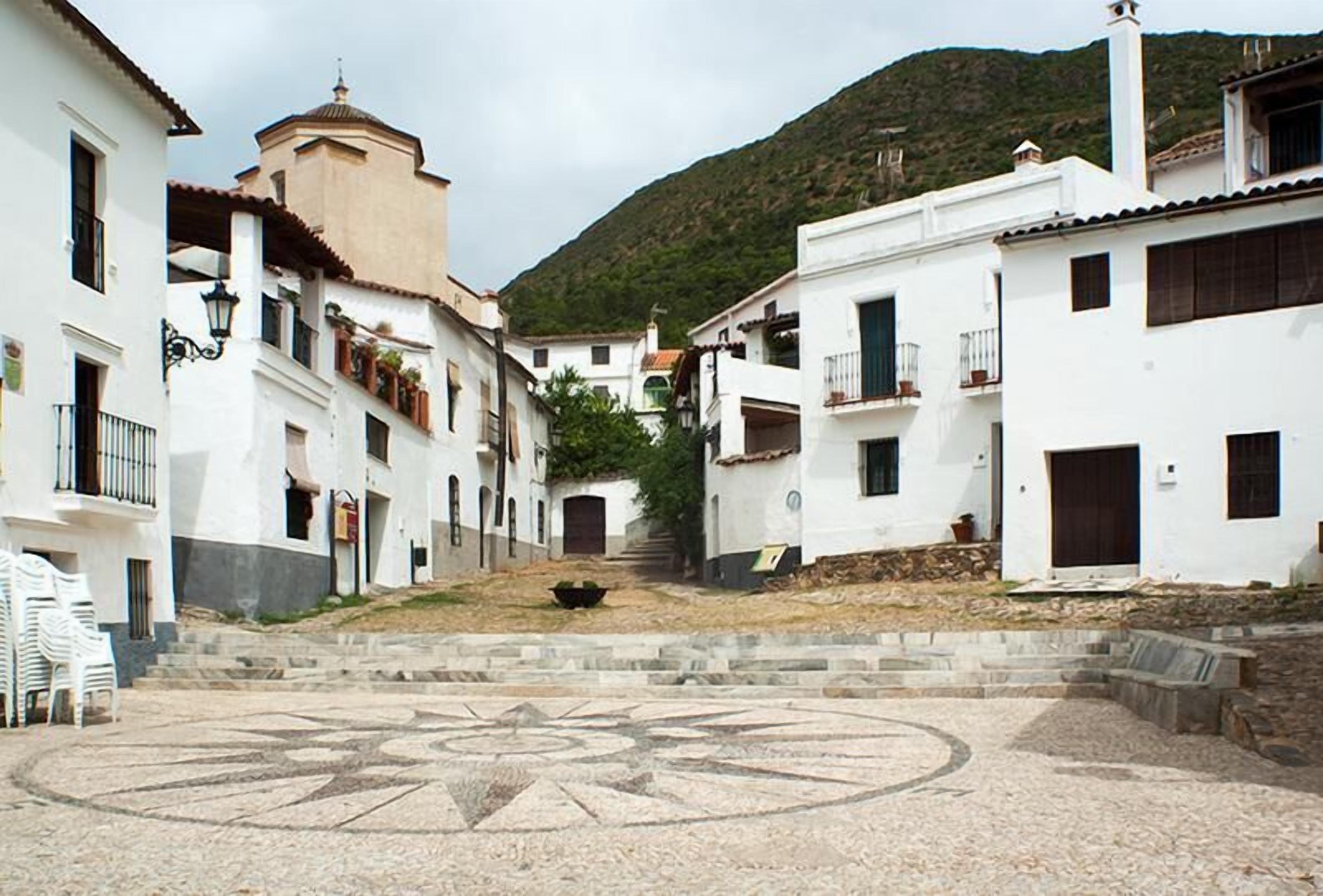 Un patio con un diseño geométrico de azulejos, rodeado de edificios blancos con balcones y una escalera central que sube a una colina.