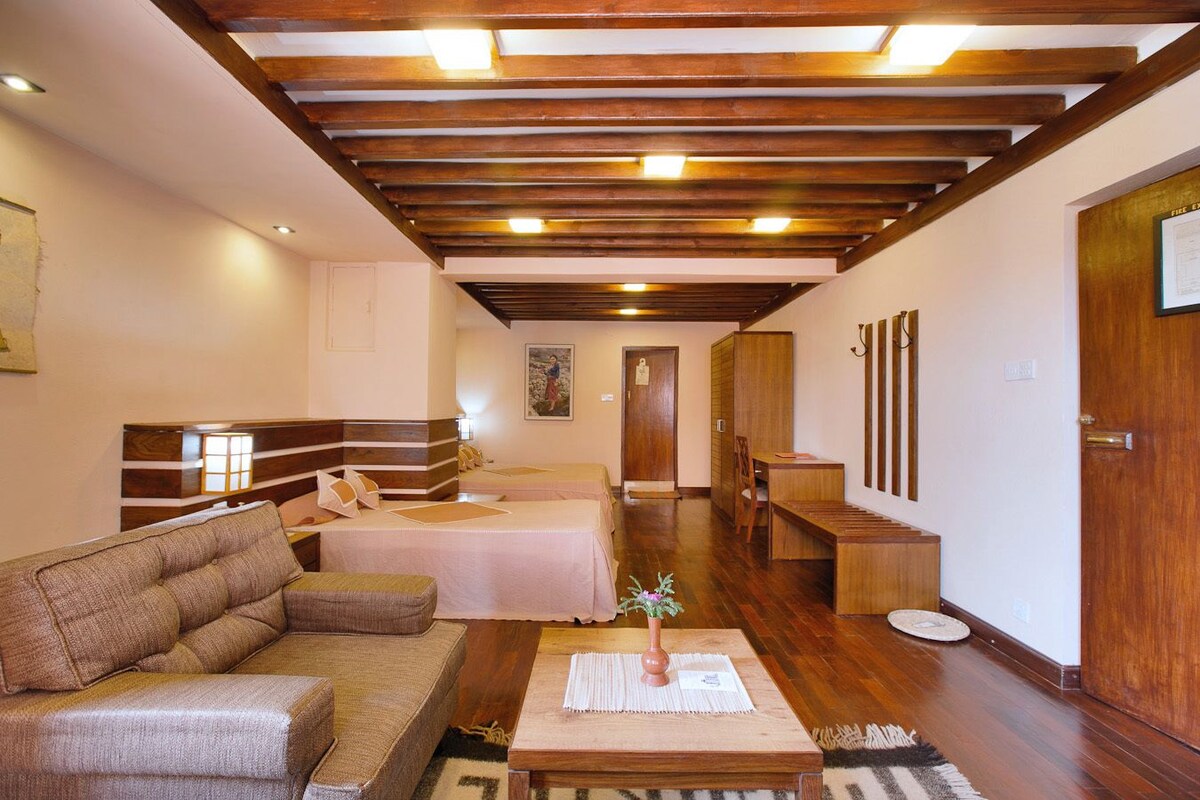 A hotel room with a wooden ceiling, a sofa, a bed, a small table with a vase, and a wooden bench.