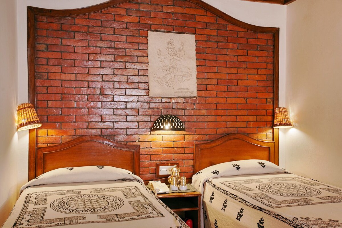 A hotel room with two beds, a brick wall, a mounted artwork, and two wall-mounted lamps.