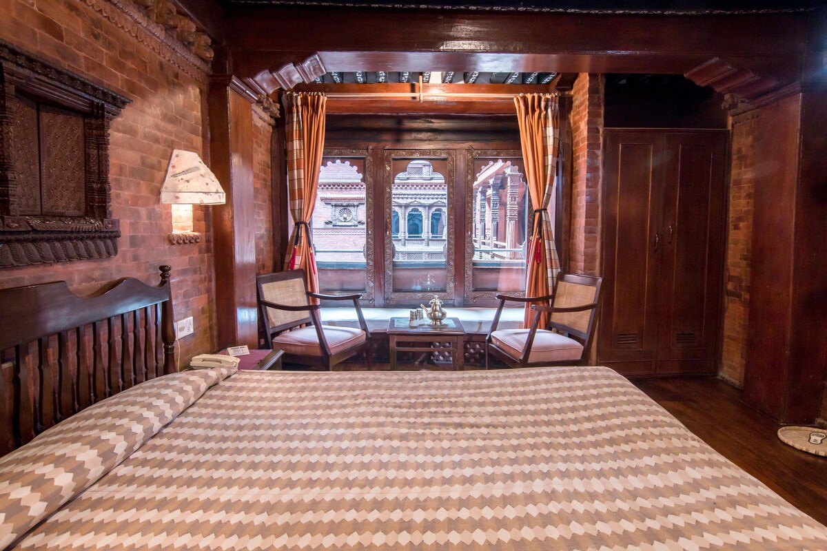 A room with a bed, two chairs, a table with a teapot, and a view of a building through a window with curtains.