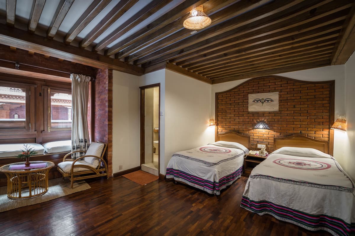 Habitación con dos camas, piso de madera, pared de ladrillo decorativa, una silla de mimbre, una mesita con una planta y una ventana con cortinas.