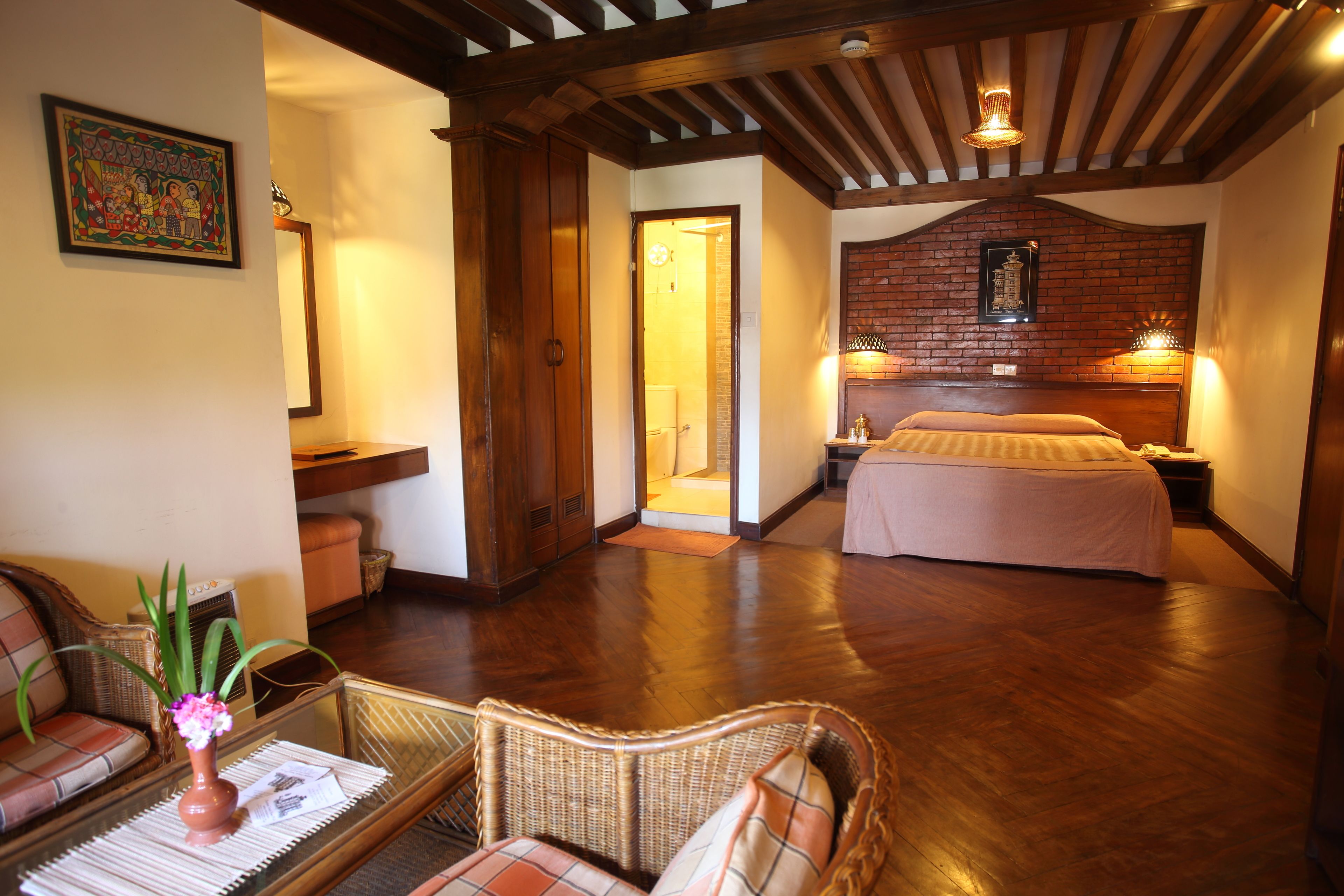 Een ruime kamer met houten vloer, een bed met hoofdeinde, een rieten stoel, een klein tafeltje met een vaas, en een deur naar een andere kamer.