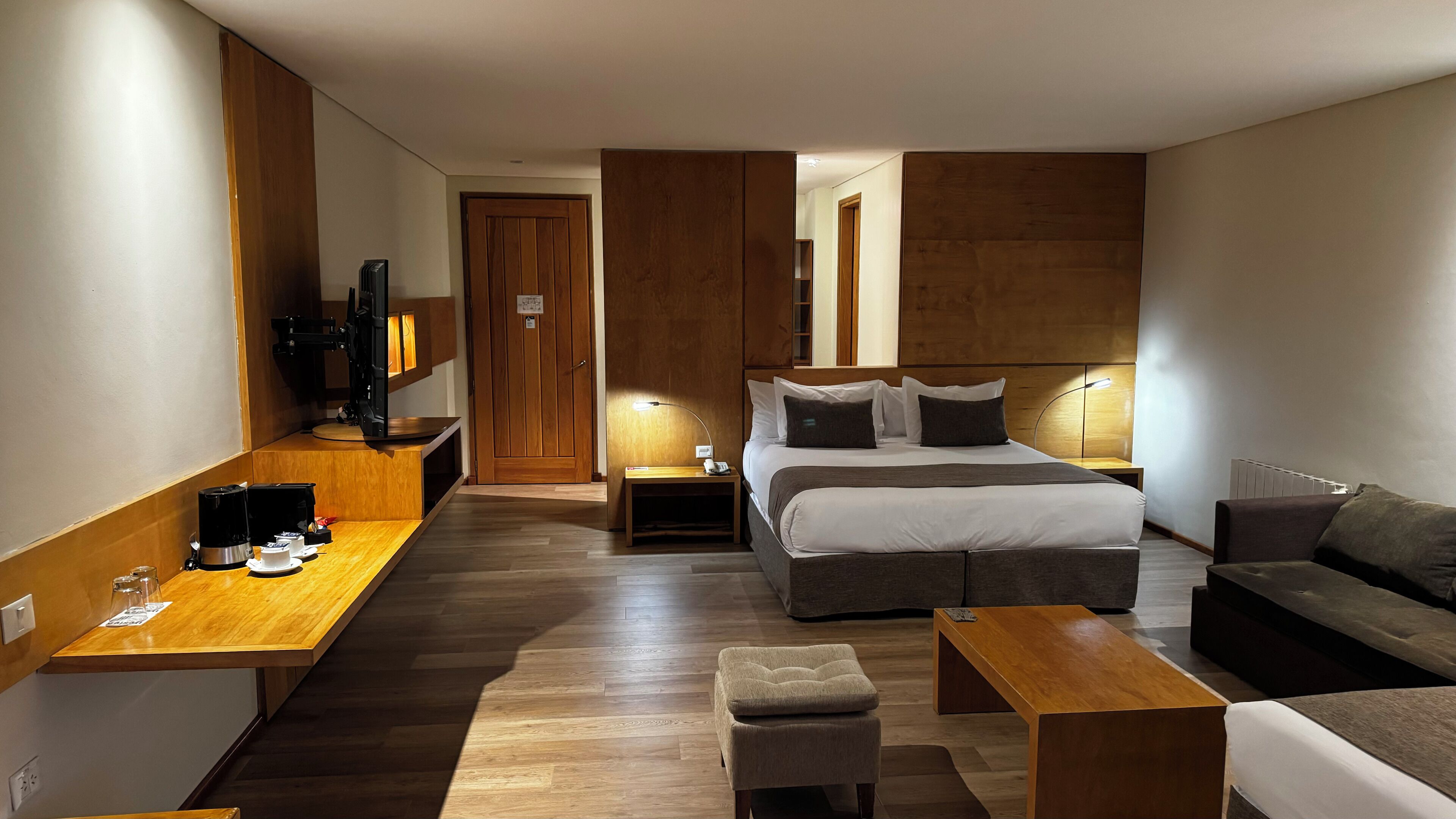 A modern hotel room with a wooden floor, a bed with white linens, a sofa, a small table, and a TV on a wooden stand.
