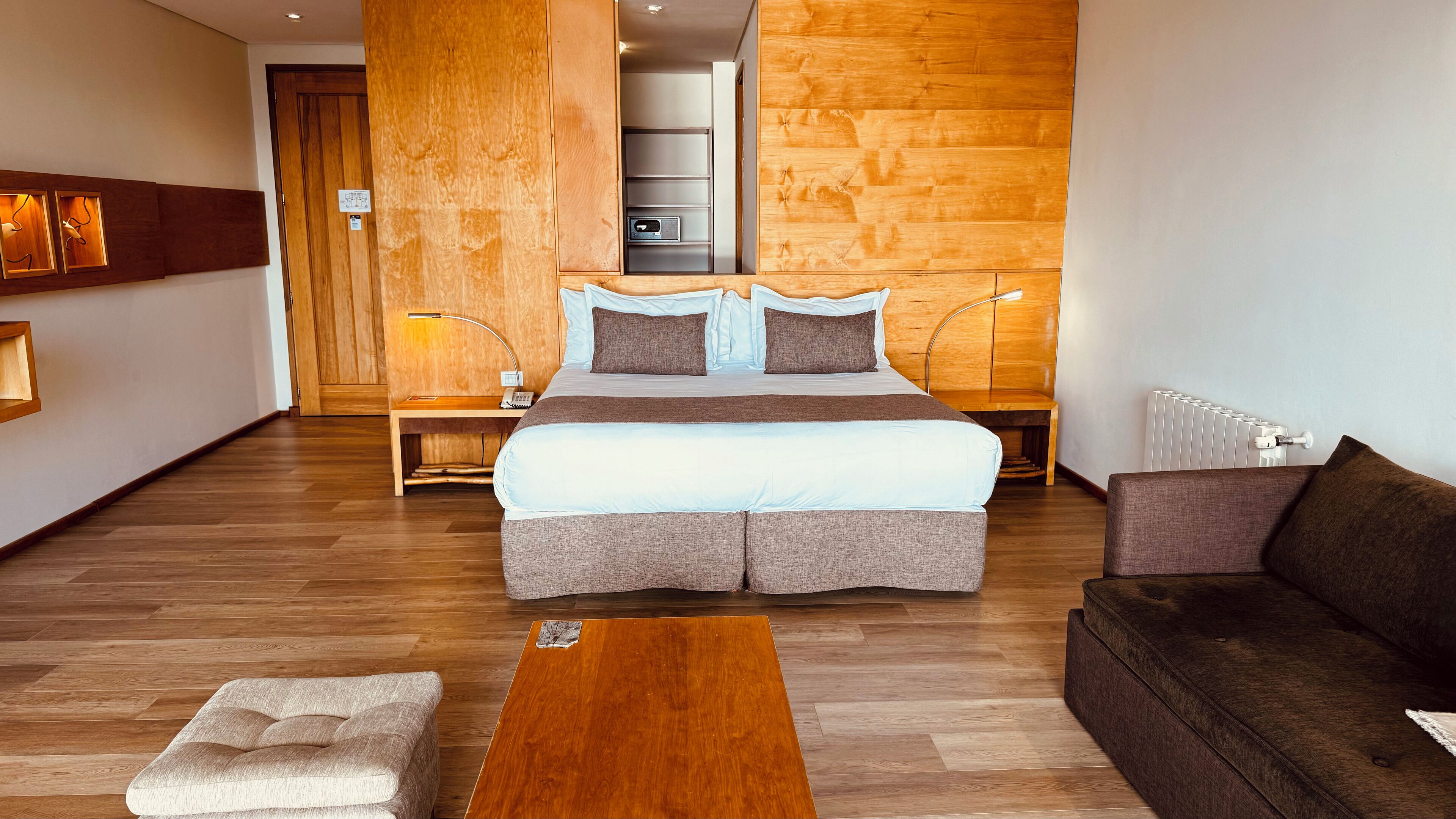 A hotel room with a large bed, a wooden headboard, a wooden floor, a sofa, a coffee table, and a nightstand with a lamp.