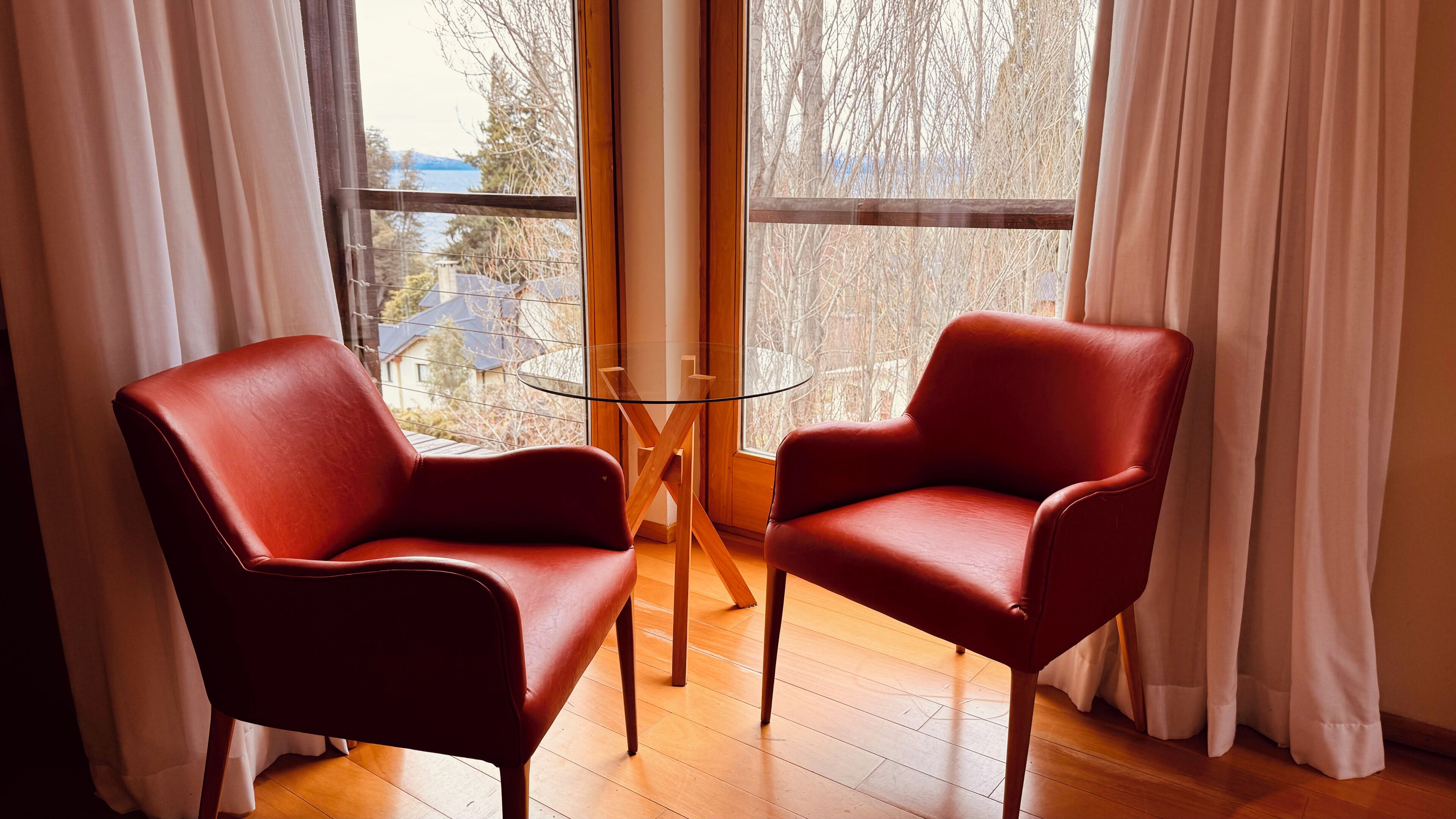 Dos sillones de cuero rojo frente a una mesa de vidrio en una habitación con suelo de madera y ventanas amplias con cortinas blancas.