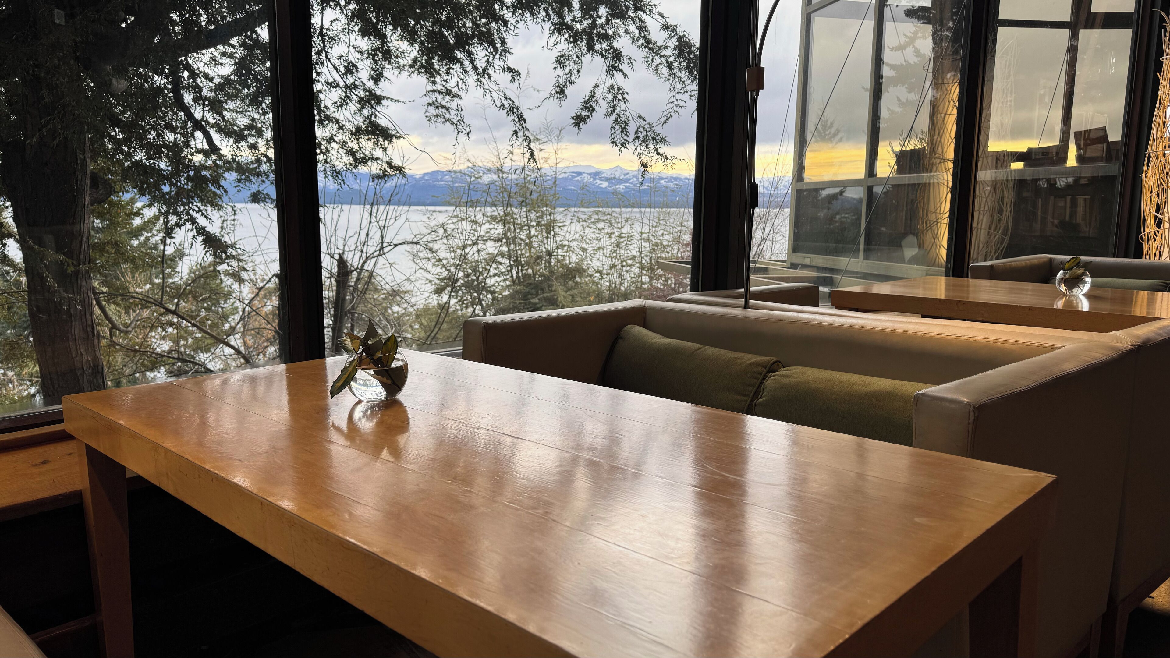 Una mesa de madera con un florero de vidrio, un banco acolchado y vistas a montañas y un lago a través de amplias ventanas.