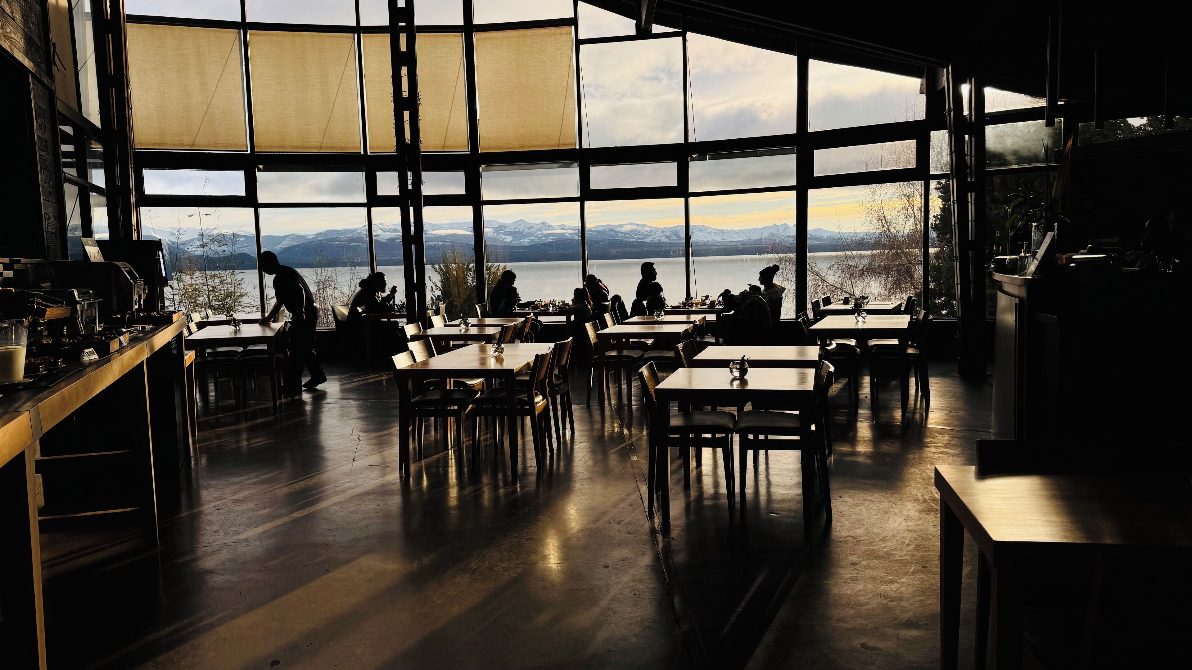 Ein Restaurant mit großen Fenstern, das einen Blick auf einen See und Berge bietet. Tische und Stühle sind für das Essen bereitgestellt.