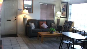 A living room with a sofa, coffee table, and chairs. There is a door with an 'EXIT' sign, a lamp, and a window with blinds. - Lodge on the River (Bullhead City)