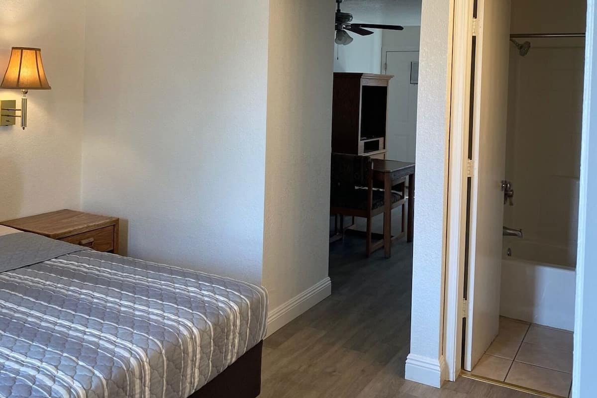 A bedroom with a bed, a desk, a lamp, and a door leading to a bathroom.