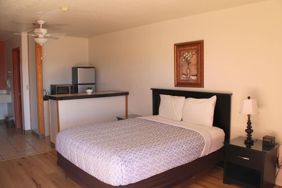 A bedroom with a bed, a nightstand with a lamp, a desk with a computer and a plant, and a painting on the wall.