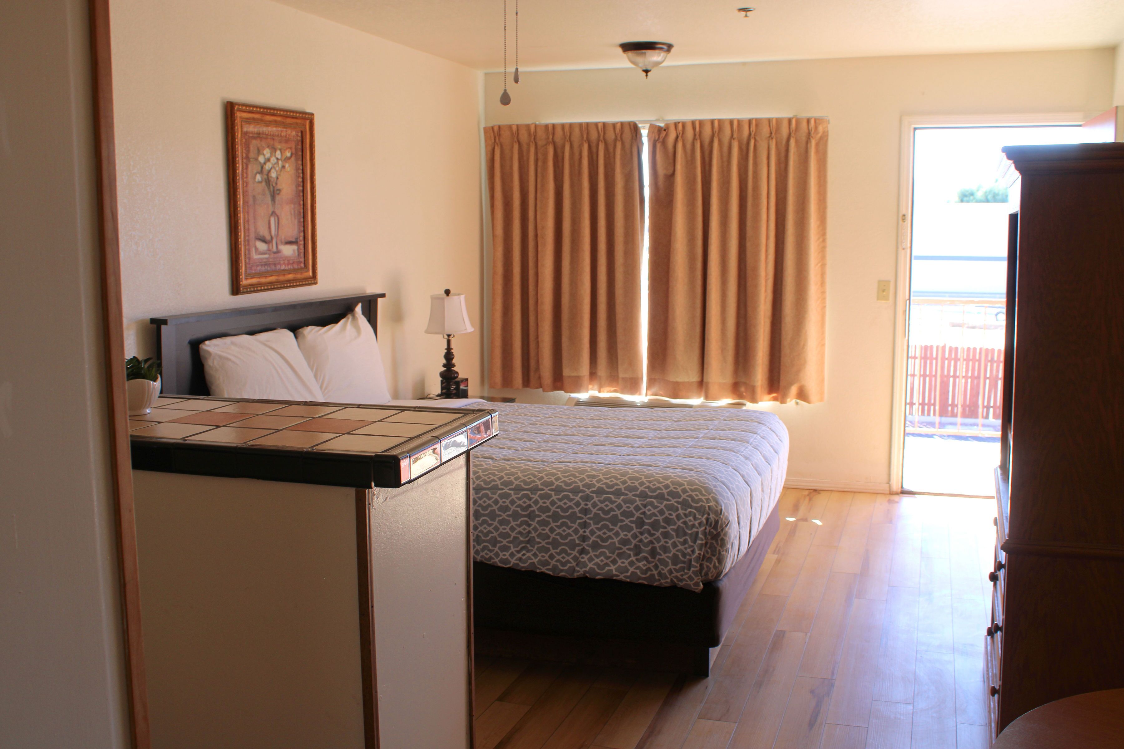 Habitación de hotel con cama, mesita de noche, lámpara, ventana con cortinas y puerta que da al exterior.