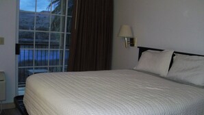 A neatly made bed with a white quilt, a bedside lamp, and a window with curtains. - Lodge on the River (Bullhead City)