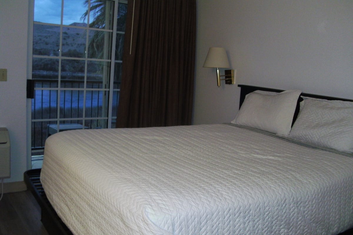A neatly made bed with a white quilt, a bedside lamp, and a window with curtains.