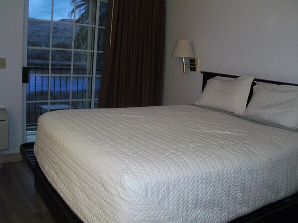 A neatly made bed with a white quilt, a bedside lamp, and a window with curtains. - Lodge on the River (Bullhead City)