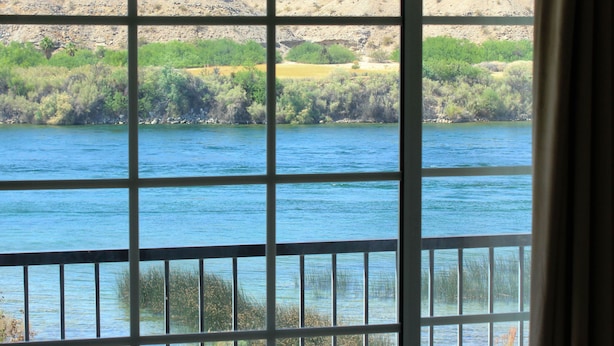 A view through a window with a metal grid, overlooking a body of water with distant hills and sparse vegetation. - Lodge on the River (Bullhead City)