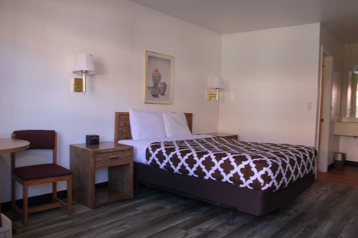 A hotel room with a bed, a wooden nightstand, a chair, and a small table.