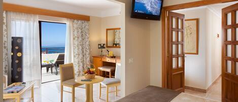 Hotellihuone, jossa on sänky, työpöytä, tuoli, hedelmillä täytetty tarjoilupöytä, televisio ja näkymä merelle.