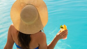 A person in a swimming pool wearing a straw hat and holding a drink with a slice of orange and mint.