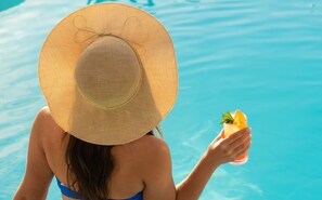 A person in a swimming pool wearing a straw hat and holding a drink with a slice of orange and mint.