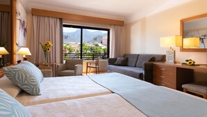 Habitación de hotel con una cama grande, un sofá, un escritorio y una silla. Se ve una ventana con vistas a montañas y palmeras.