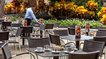 An outdoor café with wicker chairs and tables, a waiter in the background, and tropical plants.