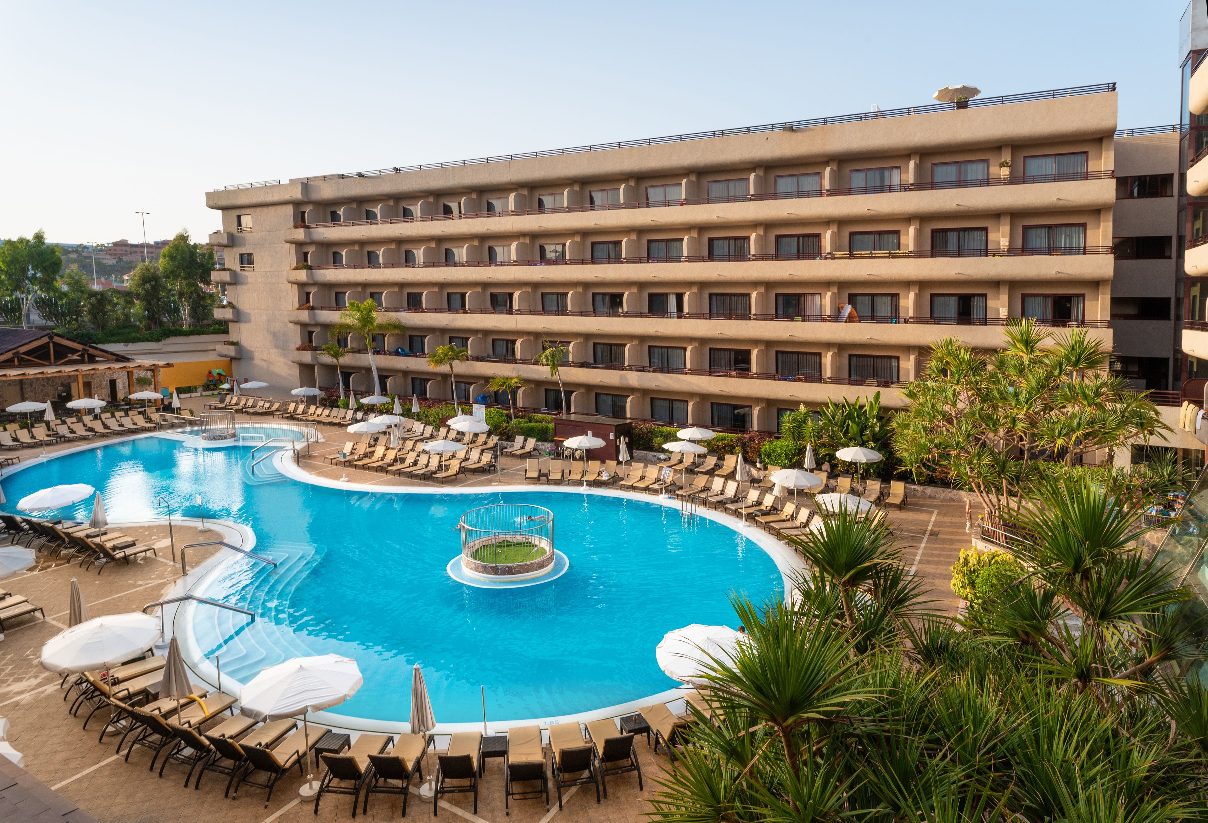Un hotel con una piscina grande rodeada de tumbonas y sombrillas, un jacuzzi pequeño y un edificio de varios pisos con balcones.