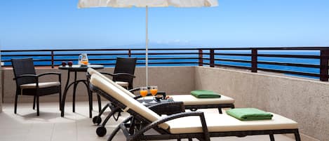 Un balcón con sillones de descanso, una mesa con frutas y bebidas, y vistas al mar.