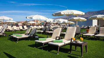 A beachside lounge area with rows of sun loungers and white umbrellas, a small table with drinks, and a view of the sea and mountains in the background.