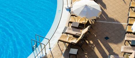 Uma área à beira da piscina com espreguiçadeiras e guarda-sóis, piscina azul cristalina e um deck revestido de azulejos.