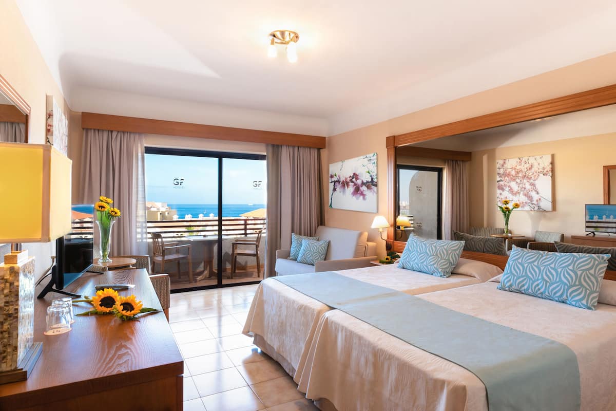 A hotel room with two beds, a desk, a chair, a TV, and a balcony with a view of the sea.
