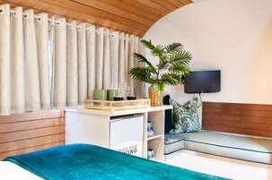 A bedroom with a bed, a bedside table with a lamp, a plant, and a window with curtains.