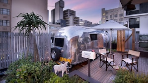 Une remorque métallique surmontée d’une enseigne indiquant « Rooftop Safari », installée sur une terrasse en bois avec des chaises et une table, le tout entouré d’une clôture en bois et de verdure.