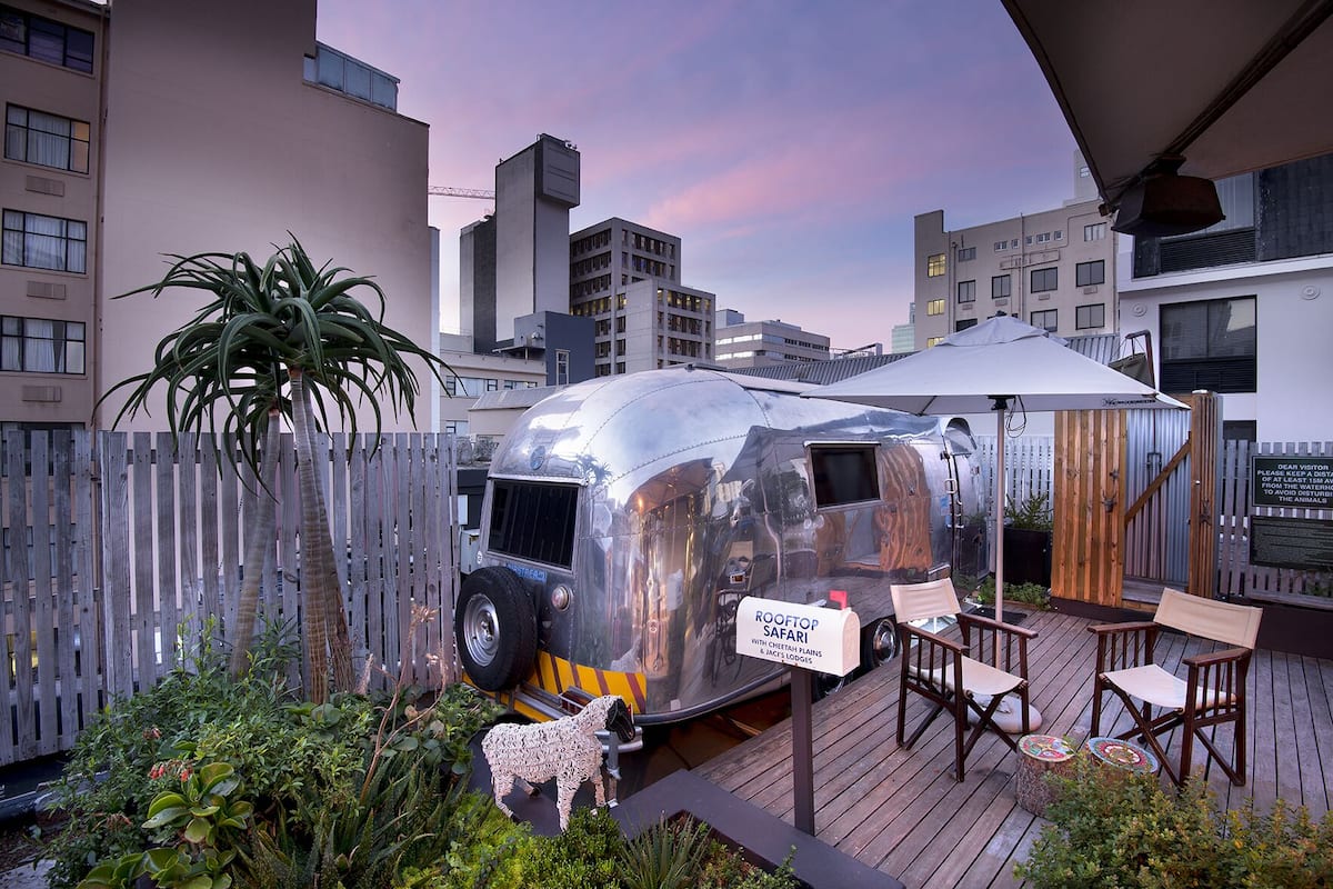 Ein metallischer Anhänger mit dem Schriftzug "Rooftop Safari" auf einer Holzdielenfläche mit Stühlen und einem Tisch, umgeben von einem Holzzaun und Grünpflanzen.