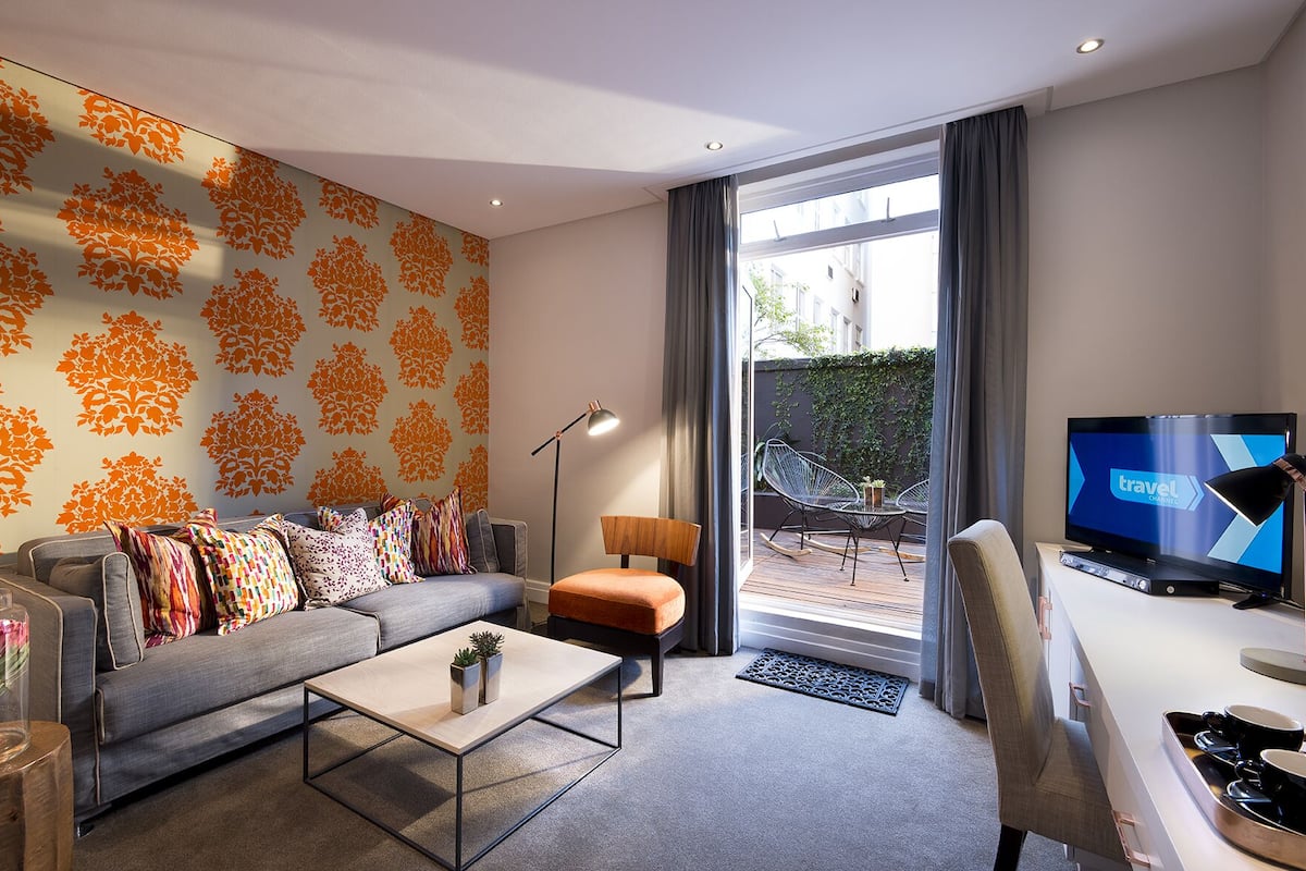 Ein modernes Wohnzimmer mit einem grauen Sofa, bunten Kissen, einem Holztisch und einem Fernseher, der das Wort 'travel' anzeigt.