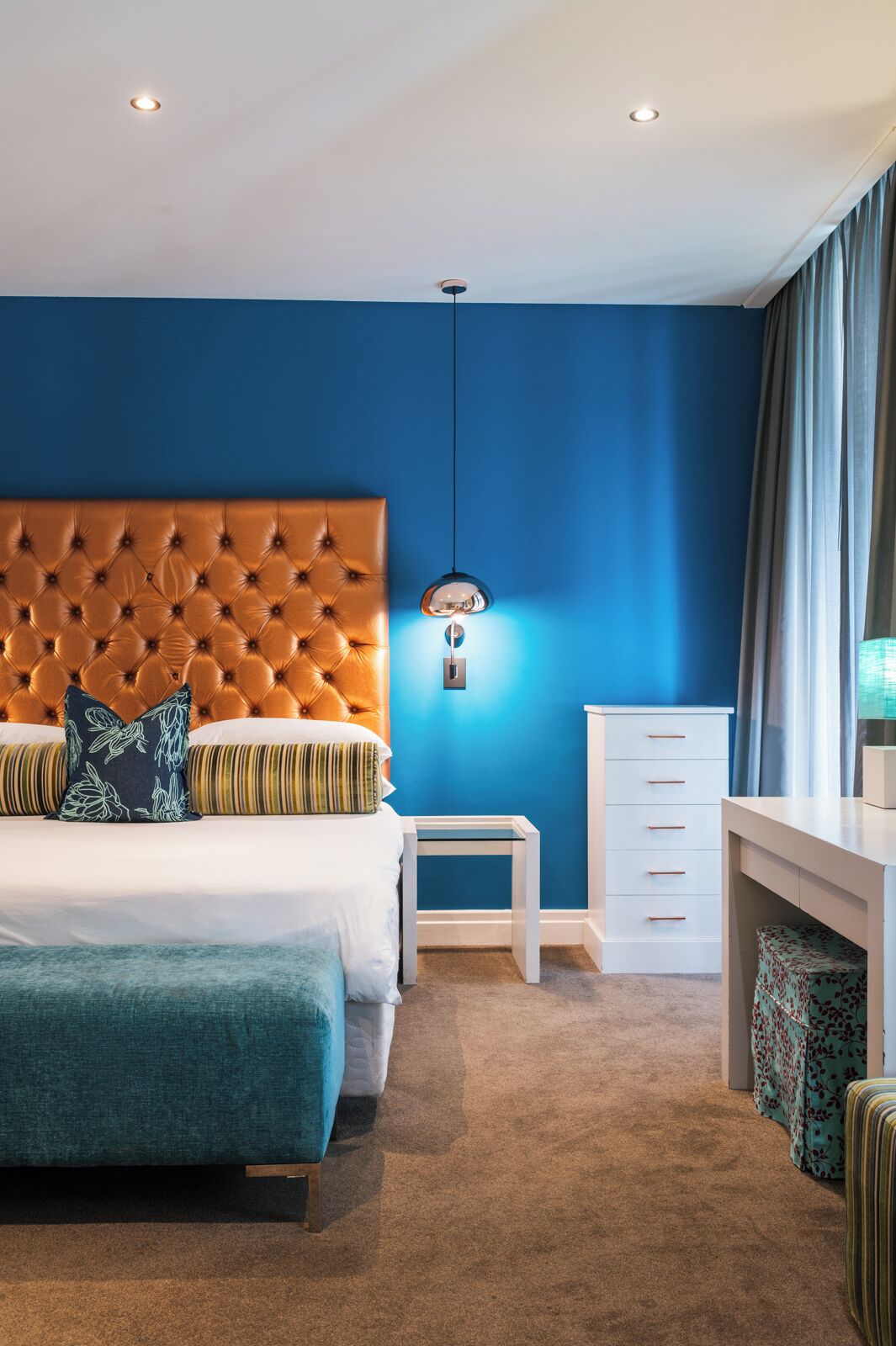 A bedroom with a tufted headboard, a bedside table, a dresser, and a window with curtains.